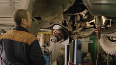 Asyalı bir adam atölyedeki asansörde kaldırılan eski bir arabanın şasesini tamir ediyor..