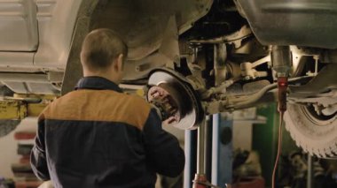 Asyalı bir adam atölyedeki asansörde kaldırılan eski bir arabanın şasesini tamir ediyor..
