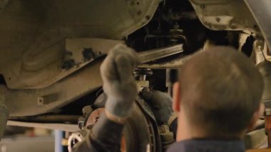 Asyalı bir adam atölyedeki asansörde kaldırılan eski bir arabanın şasesini tamir ediyor..