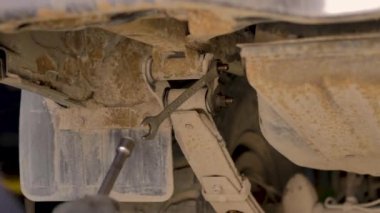 Asyalı bir adam atölyedeki asansörde kaldırılan eski bir arabanın şasesini tamir ediyor..