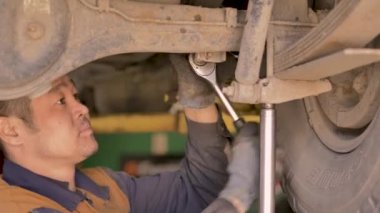 Asyalı bir adam atölyedeki asansörde kaldırılan eski bir arabanın şasesini tamir ediyor..