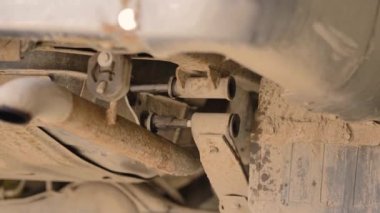 Asyalı bir adam atölyedeki asansörde kaldırılan eski bir arabanın şasesini tamir ediyor..