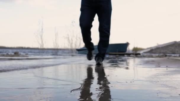 男は海岸沿いを歩くが 足だけが焦点を当てていない 背景にはボートと川があります 地球の湿った表面から反射が見える 夏の夜 — ストック動画
