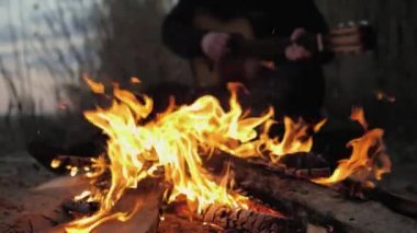 Bir adam gitar çalar. Ön planda bir yangın var. Yaz akşamı. Yavaş çekim.