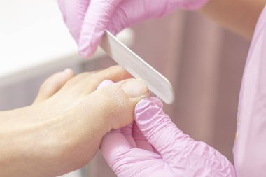 Genç bir kadın güzellik salonunda profesyonel pedikür yaptırıyor, yakın plan. Koruyucu lastik eldivenli bir pedikürcünün elleri tırnak törpüsüyle çivilenir. Profesyonel güzellik salonu. Pedikür, manikür konsepti