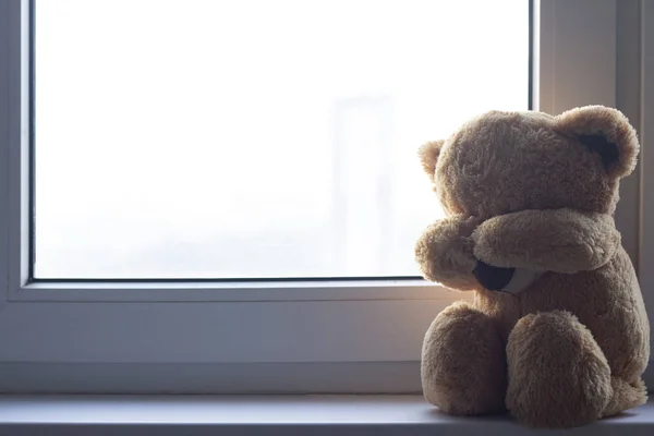 A soft toy bear cries sitting on a windowsill. There are noises in the photo, because the photo is dark.