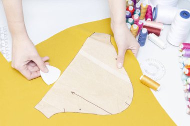 Dikiş stüdyosu. Terzi kadın kumaşın üzerindeki deseni yakın plan daire içine alıyor. Dikiş işi için bir takım ögeler: iplikler, iğneler, iğneler, makas, mezura, vs..