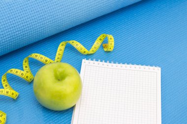 Yeşil elma, mezura, mavi renkli bir spor minderinin üzerindeki not defteri. Yaz sezonu ve plaj hazırlıkları, kilo kaybı ve spor konsepti
