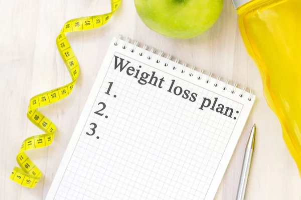 green apple, measuring tape and notebook for writing with the inscription weight loss plan on a light wooden background. preparation for the summer season and the beach, weight loss and sports concept