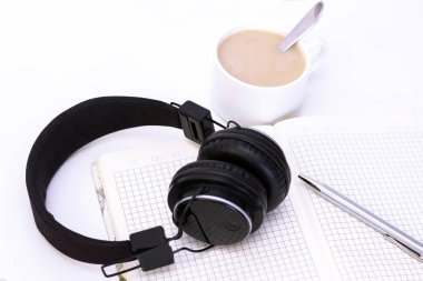 Siyah kulaklıklar ve beyaz arka planda bir fincan kahve. Masanın üzerinde kalem olan defter.