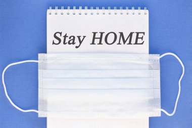 Yazıt. Evde kal. Bir kağıt parçasında, tıbbi koruyucu bir maske. Mavi bir arka planda. Coronavirus, Salgın, Evde Kalın, Güvende Kalın