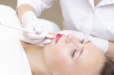 Güzellik salonunda dudaklarında kalıcı makyaj yapan genç bir kadın. Kalıcı Makyaj (Dövme) Beyaz bir dudak kalemiyle bir çizgi çizmek