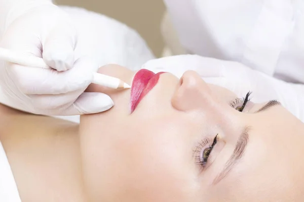 Young woman having permanent makeup on her lips at the beauticians salon. Permanent Makeup (Tattoo). drawing a contour with a white lip pencil