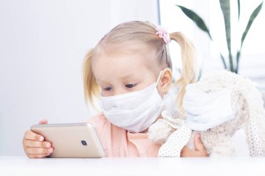 Koruyucu maskeli bir kız, oyuncak bir tavşanla cep telefonu kullanıyor, video görüşmeleri için akıllı bir telefon, akrabalarla konuşuyor, bir kız evde oturuyor, online bilgisayar webcam, video görüşmesi yapıyor..