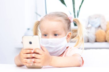 Koruyucu tıbbi maskeli bir kız cep telefonu kullanıyor, video görüşmeleri için akıllı telefon kullanıyor, akrabalarıyla konuşuyor, evde oturan bir kız, online bilgisayar kamerası, video görüşmesi yapıyor..