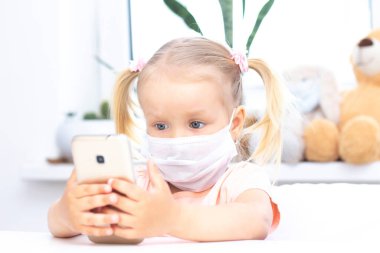 Koruyucu tıbbi maskeli bir kız cep telefonu kullanıyor, video görüşmeleri için akıllı telefon kullanıyor, akrabalarıyla konuşuyor, evde oturan bir kız, online bilgisayar kamerası, video görüşmesi yapıyor..