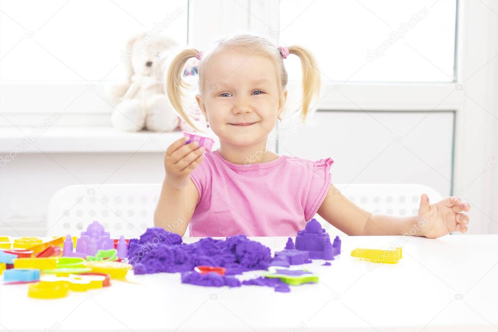 happy Girl plays kinetic sand in quarantine. Blond beautiful girl smiles and plays with purple sand on a white table.