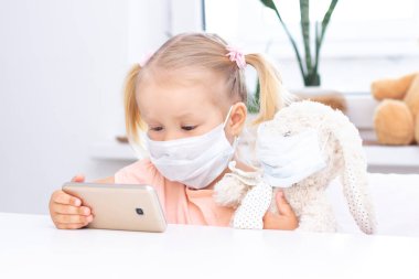 Koruyucu tıbbi maskeli bir kız cep telefonu kullanıyor, video görüşmeleri için akıllı telefon kullanıyor, akrabalarıyla konuşuyor, evde oturan bir kız, online bilgisayar kamerası, video görüşmesi yapıyor..