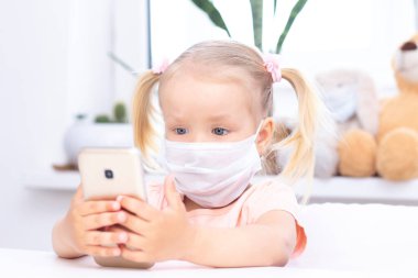 Koruyucu tıbbi maskeli bir kız cep telefonu kullanıyor, video görüşmeleri için akıllı telefon kullanıyor, akrabalarıyla konuşuyor, evde oturan bir kız, online bilgisayar kamerası, video görüşmesi yapıyor..
