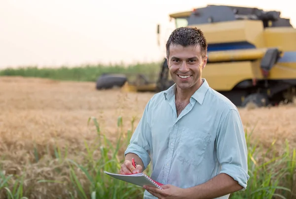 小麦畑に立つノート付きの幸せな若い農家のエンジニアと背景に働く収穫者を組み合わせる — ストック写真