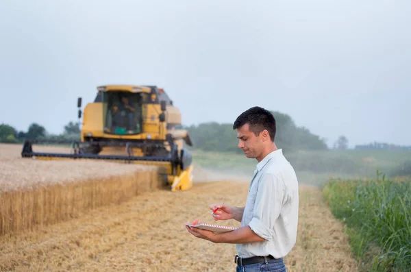 コンバインのバック グラウンドで動作中の麦畑にノートブック立って 若い農夫エンジニア — ストック写真