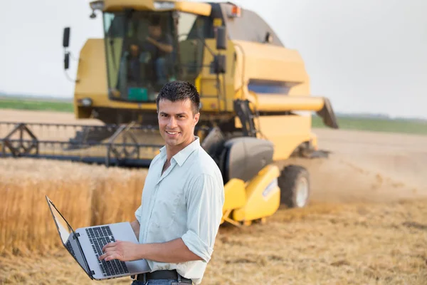 快乐年轻农夫工程师与膝上型计算机站在麦田同时联合收割机工作背景 — 图库照片