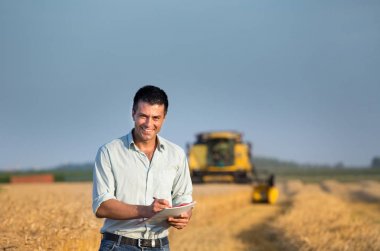 Buğday tarlasında duran ve arka planda çalışan biçerdöveri birleştiren mutlu genç çiftçi mühendisi.