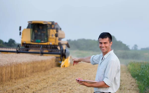 小麦畑に立つノート付きの幸せな若い農家のエンジニアと背景に働く収穫者を組み合わせる — ストック写真