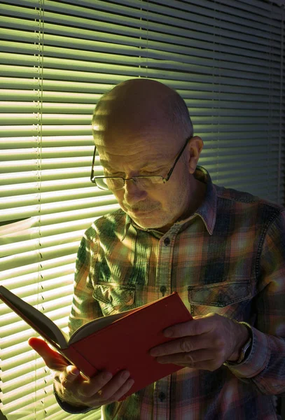 Az öregember könyvet olvas az ablak mellett. — Stock Fotó