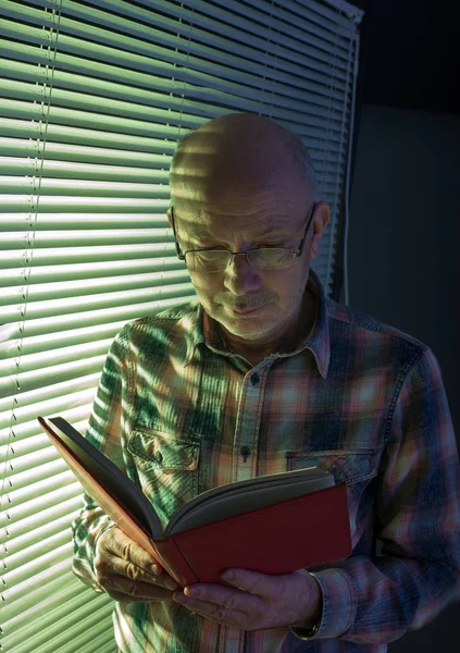 Oude man leest boek naast raam — Stockfoto