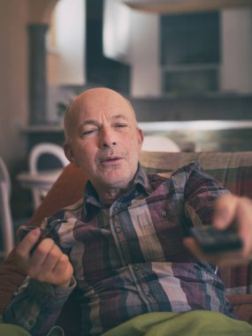 Olgun bir adam evdeki koltukta oturup uzaktan kumandayı tutuyor, pipo içiyor ve televizyon seyrediyor.