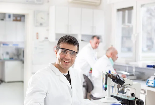 研究室にいる同僚と顕微鏡の横に座って笑っているハンサムな生物学者の肖像画 — ストック写真