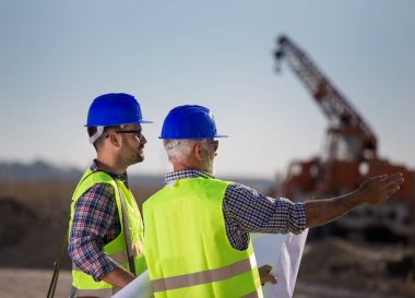 İki deneyimli mühendis planlara bakıyor ve arkaplanda metal vinçle inşaat alanında konuşuyorlar.