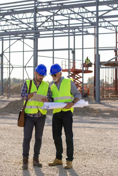 İnşaat alanındaki tablete bakan iki mühendis ve arka planda metal yapı var.