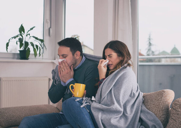 Young couple sitting on sofa covered with blanket, sneezing and blowing nose. Cold and flu season 