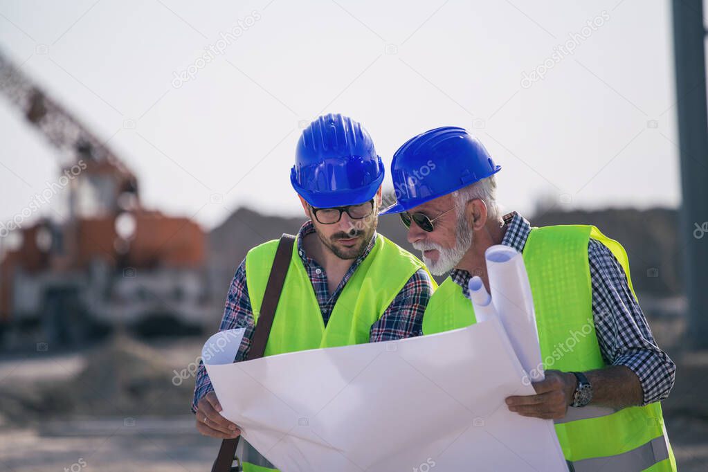 Two experienced engineers looking at blueprints and talking at building site with metal crane in background