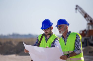 İki deneyimli mühendis planlara bakıyor ve arkaplanda metal vinçle inşaat alanında konuşuyorlar.