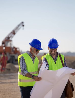 İki deneyimli mühendis planlara bakıyor ve arkaplanda metal vinçle inşaat alanında konuşuyorlar.
