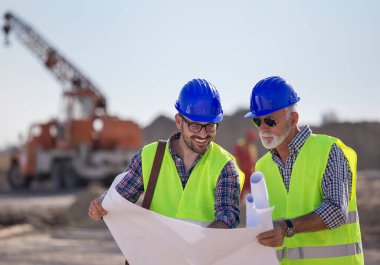 İki deneyimli mühendis planlara bakıyor ve arkaplanda metal vinçle inşaat alanında konuşuyorlar.