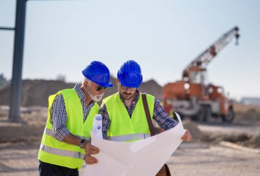 İki deneyimli mühendis planlara bakıyor ve arkaplanda metal vinçle inşaat alanında konuşuyorlar.