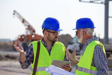 İnşaat alanında konuşan tabletli ve planlı iki mühendis ve arka planda metal yapı var.