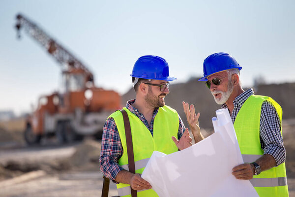 Two experienced engineers looking at blueprints and talking at building site with metal crane in background