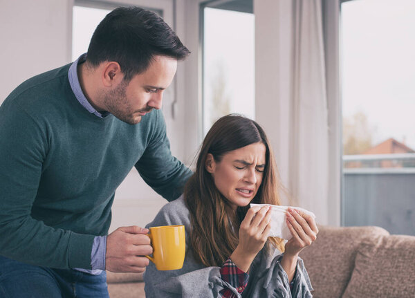 Boyfriend bringing tea and taking care of girl having flu. Sitting on sofa at home covered with blanket and sneezing