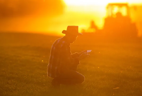 Bauer Hockt Bei Sonnenuntergang Vor Traktor Und Schaut Auf Tablet — Stockfoto