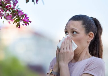 Genç ve güzel bir kadın çiçek açan ağacın önünde burnunu siliyor. Bahar alerjisi konsepti