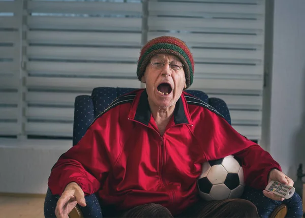 兴奋的老人坐在家里的扶手椅上 拿着足球 看电视上的足球比赛 — 图库照片