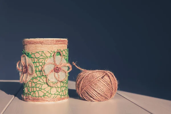 A skein of jute rope and a metal jar decorated with burlap and a flower of thread and buttons, a glass for brushes and tool accessories. The concept of needlework and manual creativity.