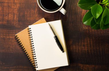 Boş sayfaları olan açık okul defterinin en üst görüntüsü. Ahşap masa arkasında Pen, Bitki ve Kahve Bardağı. Kopya alanı ile iş, ofis veya eğitim kavramı.