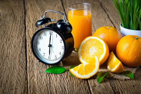 Close-up of Retro clock vintage with glass orange juice and fresh fruits ripe cut half, slice with green leaves on old wood table. Breakfast food or beverage for morning. Organic and healthy Concept.