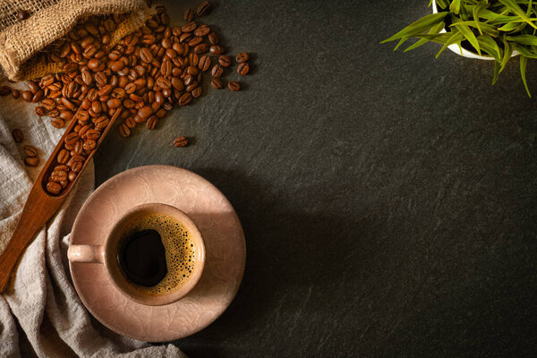 Close-up of Black hot coffee for morning with milk foam in old pink ceramic cup and napkin with coffee beans roasted in sack on dark table stone background. Top view, flat lay with copy space.